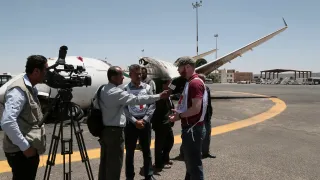 A journalist stands on the tarmac at Sana’a International Airport being interviewed by a local television crew. A passenger aircraft is parked in the background under a clear sky, with airport buildings visible beyond the runway.